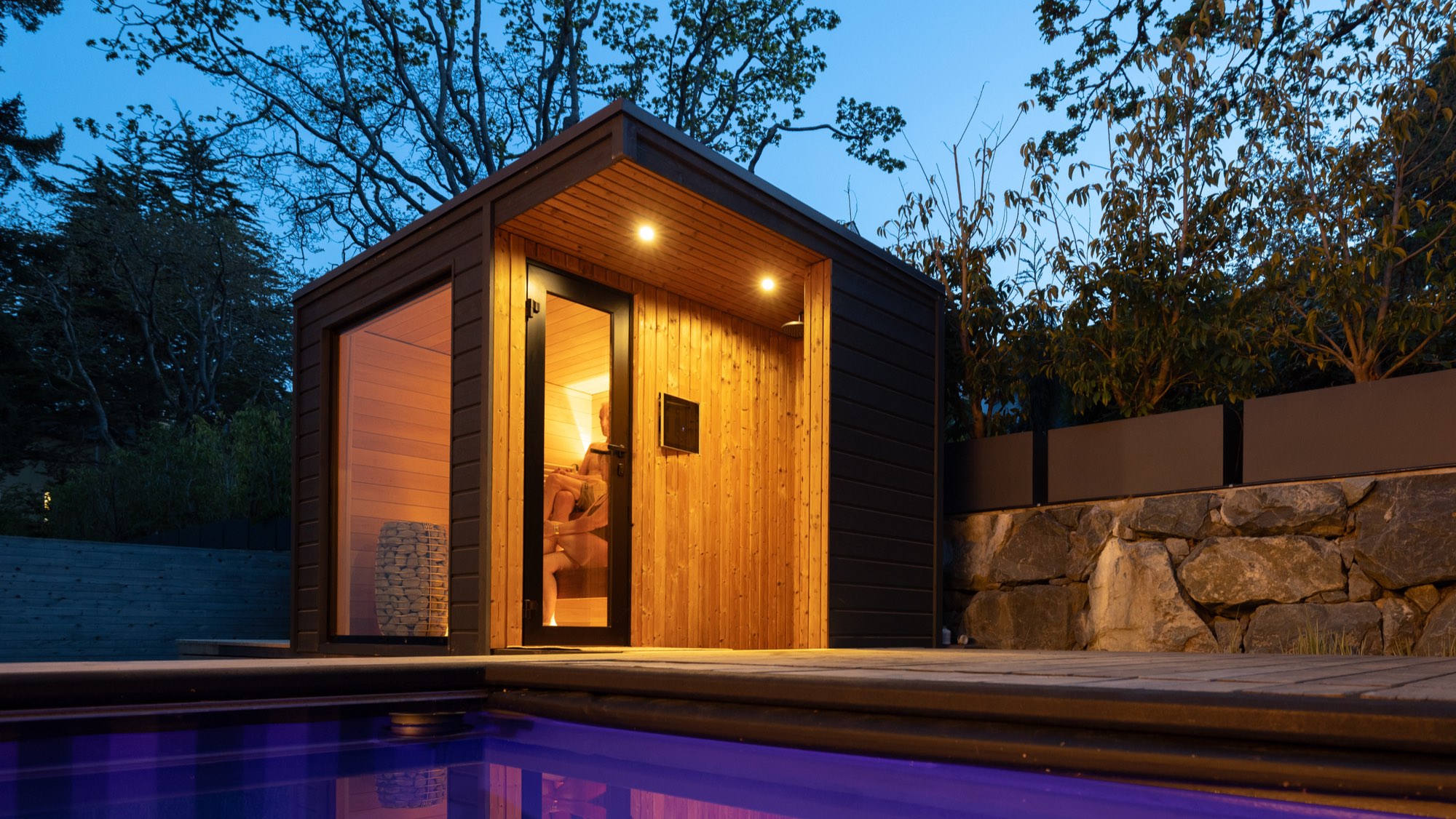 A modern outdoor sauna glowing beside water at dusk